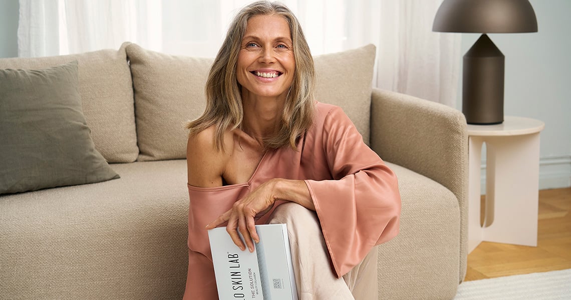 Glad kvinna sitter framför soffa och håller i en förpackning av kollagenpulver från Oslo Skin Lab